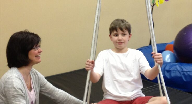 A child sitting cross-legged on a therapy swing with a therapist nearby.