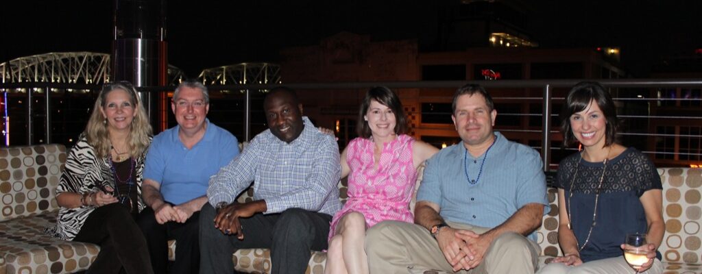 A group of six people sitting on a couch outdoors at night, smiling.