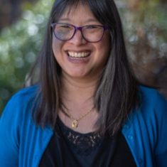 Smiling woman with glasses in a blue cardigan outdoors.
